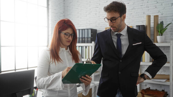 Hispanic man and woman executives collaborate in a modern office setting, reviewing documents and discussing business strategies indoors.