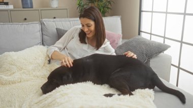 Genç İspanyol kadın labrador köpeğiyle rahat bir kanepede şık dekorlu parlak bir şehir evinde dinleniyor..