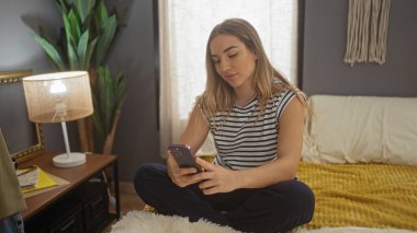 Genç, sarışın, beyaz bir kadın yatak odasında rahat bir ev ortamında akıllı telefon kullanıyor..