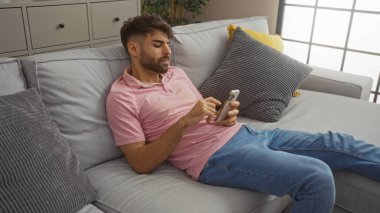 Sakallı genç adam, dairesindeki modern oturma odasındaki koltukta uzanırken akıllı telefon kullanıyor.