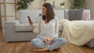 Genç, İspanyol bir kadın rahat bir oturma odasında yerde oturuyor, elinde kahve fincanı tutarken telefonuna gülümsüyor..