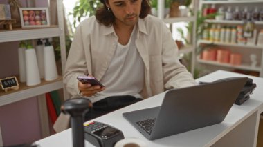 Uzun saçlı genç adam ev dekorasyonu mağazasında telefon ve laptop kullanıyor. İçi şişe ve süslemelerle dolu.