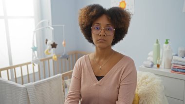 Yatak odasında beşiğin yanında kıvırcık saçlı, gözlüklü ve pembe süveterli, kapalı bir odada oturan Afro-Amerikalı genç bir kadın..