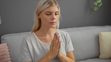 Güzel sarışın kadın huzur dolu bir oturma odasında meditasyon yapıyor. Daire ortamında rahatlık ve sakinlik gösteriyor.