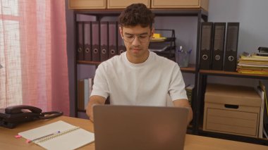 Genç bir İspanyol, bir ofiste dizüstü bilgisayarla çalışıyor. Raflarda klasörler ve organize evrak işleri var..