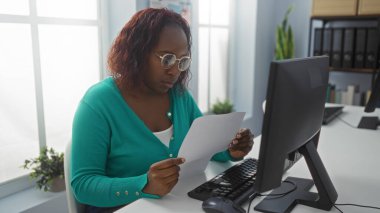 Ofis ortamında çalışan Afrikalı Amerikalı bir kadın masasında bilgisayarlı ve arka planda bitkileri olan bir belgeyi okumaya odaklanmış..