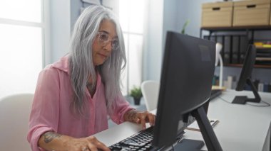 Pembe gömlekli yaşlı kadın parlak ofiste masaüstü bilgisayarında çalışıyor.