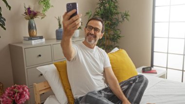 Canlı dekor ve bitkilerle dolu rahat bir yatak odasında otururken evde selfie çeken olgun İspanyol bir adam.