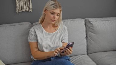 Sarı saçlı genç bir kadın, rahat bir oturma odasında gri bir koltukta otururken akıllı telefon kullanıyor..