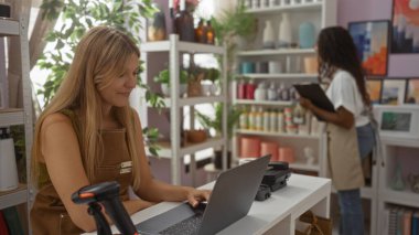Kadınlar bir mağazada çalışırken, bir kadın tezgahta dizüstü bilgisayar kullanırken, diğeri de çeşitli dekoratif eşyalarla çevrili bir panoda envanterleri kontrol ediyor.