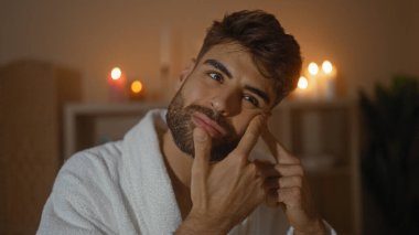 Young man with beard in spa beauty center removing pimple indoors with candles in the background creating a relaxing wellness ambiance clipart