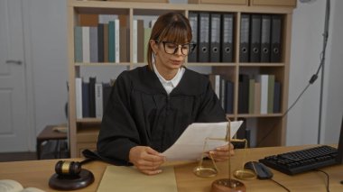 Kadın yargıç bir ofiste çalışıyor, masasında hukuk kitapları ve hukuki sembollerle çevrili belgeler okuyor, örneğin tokmak ve adalet terazisi..