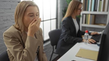 Meslektaşı bilgisayar işine odaklanırken, bir kadın modern ofiste esniyor. İki sarışın çalışanıyla birlikte yorgunluk ve profesyonel çevreyi resmediyor..