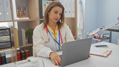 Veteriner kliniğinde çalışan çekici sarışın kadın, laptopuna odaklanmış, etrafı kitaplarla ve tıbbi aletlerle çevrili, parlak, düzenli bir çalışma alanında..