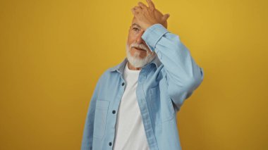 Mavi tişörtlü, sakallı, beyaz tişörtlü, sarı arka planda yalnız başına duran yakışıklı, gri saçlı bir adam..