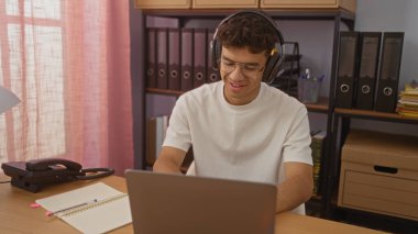 Ofisteki kulaklık ve gözlük takan genç İspanyol adam iyi organize edilmiş bir odada dizüstü bilgisayarla ve telefonla çalışıyor..