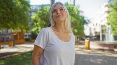 Beyaz tişörtlü çekici sarışın kadın güneşli bir günde arka planda yemyeşil ve şehir binaları olan şehir parkında güvenle gülümsüyor..