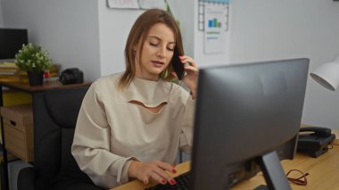 Ofiste çalışan sarışın bir kadın, bilgisayar başında çalışırken konuşuyor. Etrafı ofis malzemeleriyle çevrili, modern bir iş ortamında..