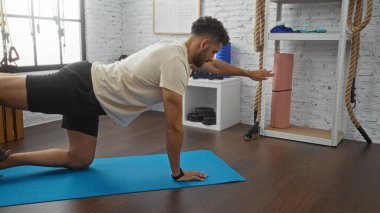 Arka planda ipler ve ekipmanlarla mavi bir paspasın üzerinde egzersiz yapan İspanyol genç adam..