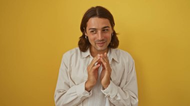 Uzun saçlı, beyaz tişörtlü, kendine güvenen ve çekici bir ifadeyle izole edilmiş sarı arka planı olan İspanyol bir genç..