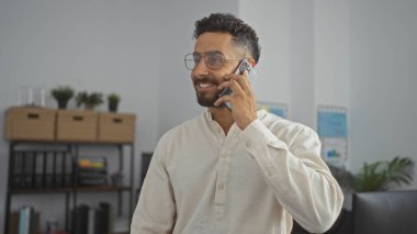 Ofiste telefonla konuşan yakışıklı İspanyol bir adam, modern kapalı bir iş yerinde güvenle gülümsüyor..