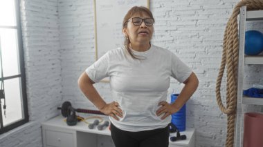 Güzel, orta yaşlı İspanyol bir kadın spor salonunda duruyor, elleri kalçasında, spor aletleri ve beyaz tuğlalarla çevrili, kapalı bir spor merkezinde canlı bir anı yakalıyor..