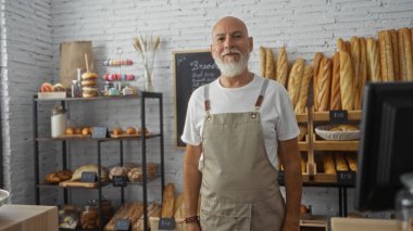 Orta yaşlı, gri saçlı ve sakallı bir adam pastanenin içinde duruyor. Arkasında ekmek ve hamur işleri rafları var..