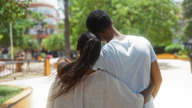 Anne ve oğlu, yeşilliklerle çevrili bir şehir parkında yürürken şefkatle kucaklaşıyorlar.