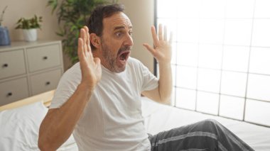 Yatakta oturan, şaşırmış yüzlü orta yaşlı bir adam.