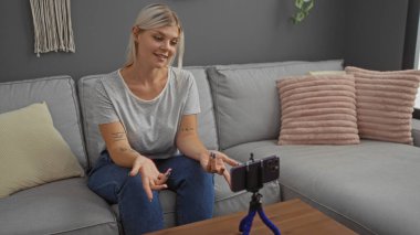 Sarı saçlı genç bir kadın evdeki bir kanepede oturuyor, oturma odasında kendini kaydediyor, akıllı bir telefonla tripoda oturuyor, gülümsüyor ve elleriyle işaret ediyor..