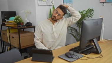 Genç İspanyol bir adam ofis çalışma odasında bilgisayarla geziyor. Çekici ve rahat görünüyor. Etrafı bitki ve ofis malzemeleriyle çevrili..