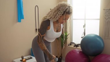 Evin içinde spor yapan bir kadın. Geniş, aydınlık bir spor salonunda, spor aletleri ve bitkilerle birlikte dambıllar kullanıyor..