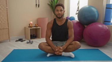 Spor salonundaki genç adam yoga minderinin üzerinde otururken gülümsüyor ve selam veriyor.