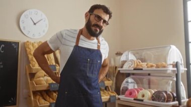 Pasta dükkanında sakallı genç bir adam. Ekmek ve ekmek sergileniyor. Gözlük ve önlük takıyor.