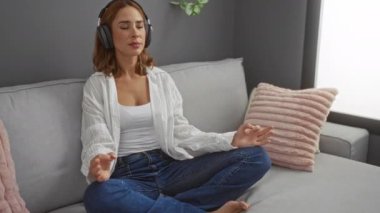 Oturma odasında kulaklıkla meditasyon yapan genç bir kadın. Kanepede bağdaş kurmuş, gözleri kapalı oturuyor. Huzurlu bir anın tadını çıkarıyor.