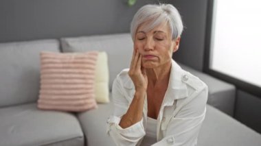 Gri duvarları ve evinde pembe bir yastıkla süslenmiş rahat bir kanepesi olan modern bir oturma odasında oturan yaşlı bir kadın diş ağrısı çekiyor..