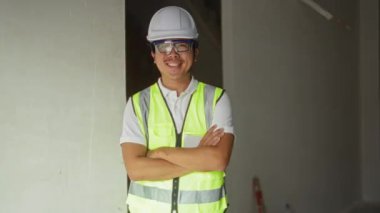 Güvenlik yeleği giyen ve kasketli genç adam kollarını kavuşturup inşaat alanında gülümsüyor.