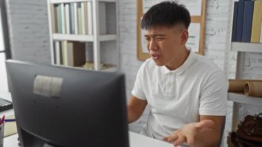 Ofis masasında beyaz gömlekli genç bir adam, arka planda raflar ve kitaplarla modern bir ofis kurulumunda otururken kapalı alandaki bilgisayar ekranına tepki veriyor.