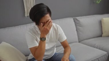 Kafasını ve gözlüklerini tutarken stresli görünen genç bir adam oturma odasında arka planda gri bir kanepede oturuyor.