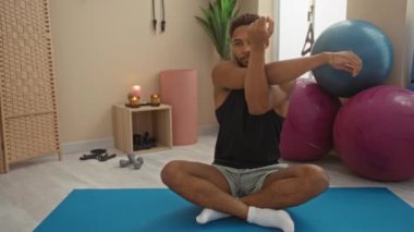 Spor salonundaki mavi bir paspasın üzerinde oturan genç adam. Egzersiz topu, mumlar ve egzersiz aletleriyle spor yaparken esneme egzersizleri yapıyor.