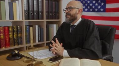 Amerikan bayrağı, hukuk kitapları ve adalet terazisiyle ofiste konuşan sakallı bir erkek yargıç.