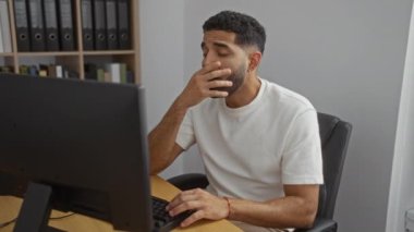 Evdeki yorgun genç adam ofis masasında dinleniyor. Bilgisayarın önünde. İş yerinde beyaz gömlek giyiyor ve modern iç mekanda ahşap masa var.