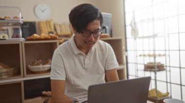 Genç adam pastanede dizüstü bilgisayarda çalışıyor. Arka planda çeşitli ekmek ve hamur işleri var.