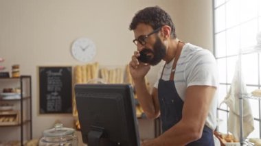 Pastanede çalışan genç bir adam, ekmek ve hamur işleriyle dolu bir tezgahın yanında önlük giyerken akıllı telefondan konuşuyor.