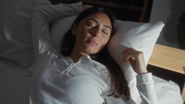 Loş ışıklı bir yatak odasında, bir yatakta huzur içinde uyuyan bir kadın, bir ev ortamında gece uykusunun sükunetini gösteriyor..