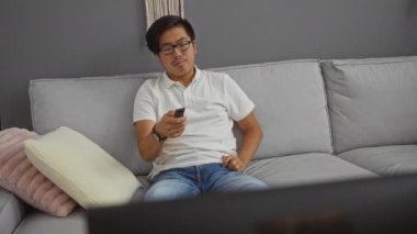 Tarafsız bir dekorla modern bir oturma odasında dinlenen genç bir adam, elinde uzaktan kumandayla evde rahat gri bir koltukta oturuyor.