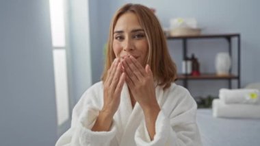 Kaplıca odasında gülümseyen, raflı beyaz bir bornoz giyen ve arka planda kaplıca eşyaları olan genç bir kadın.