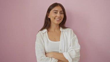 Pembe arka planda duran genç bir kadın, kollarını kavuşturmuş sıcak bir şekilde gülümsüyor, beyaz bir elbise giyiyor.