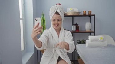 Kadın spa odasında kafasında bornoz ve havluyla selfie çekiyor, etrafı mumlar ve havlular gibi sıhhatle çevrili, rahatlama ve güzellik gösterisi yapıyor.
