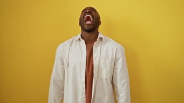 Kızgın Afrikalı Amerikalı adam sarı arka planda tek başına duruyor, tişört giyiyor, öfke ve hayal kırıklığını dile getiriyor, yüksek sesle bağırıyor ve agresif bir öfkeyle bağırıyor.